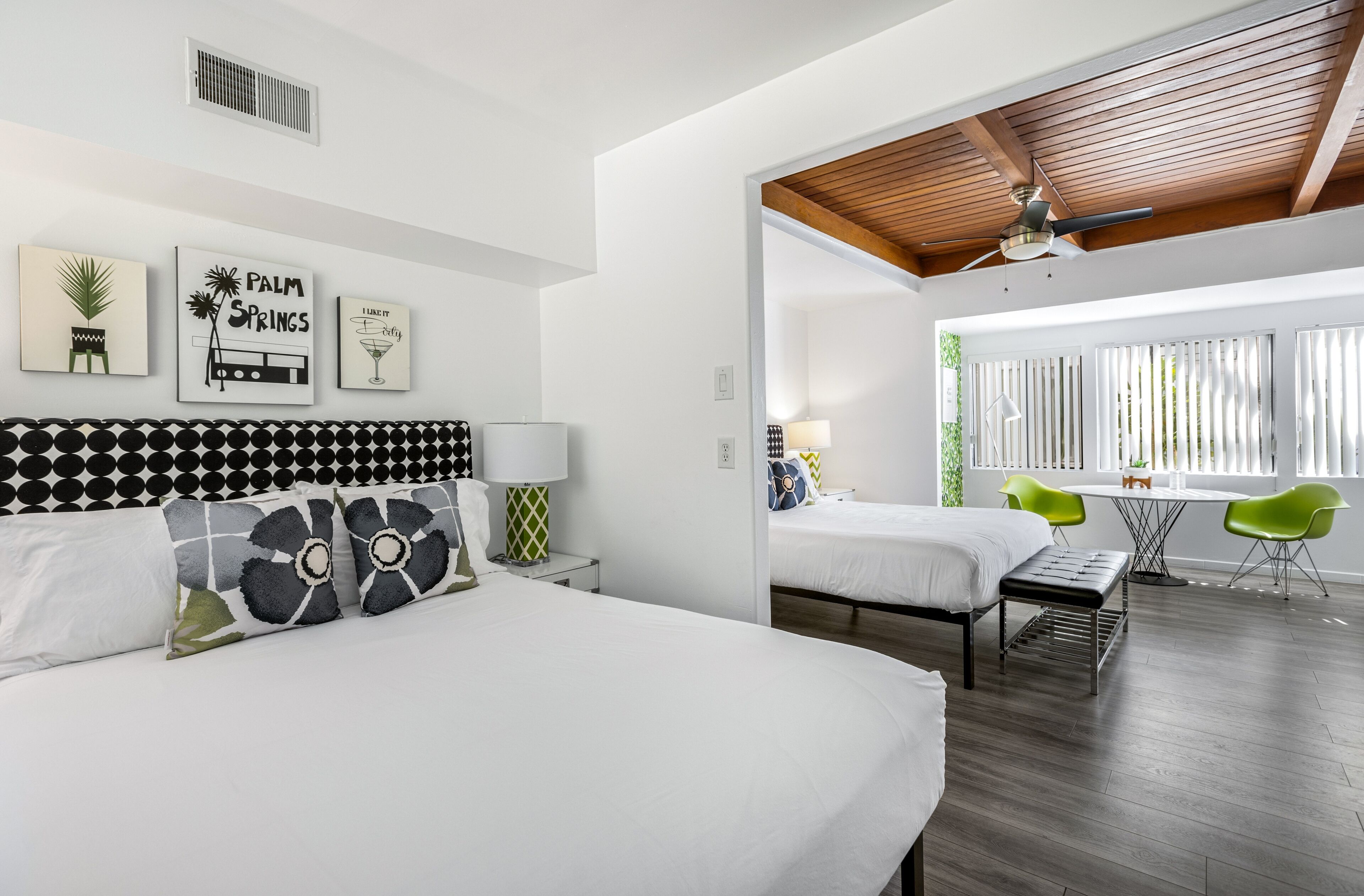 A modern hotel room with two beds, a dining area with green chairs, and a ceiling fan.