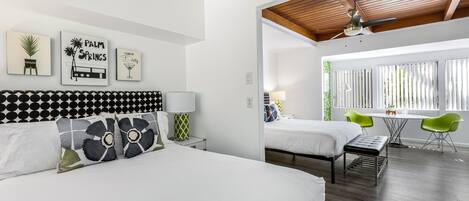 A modern hotel room with two beds, a dining area with green chairs, and a ceiling fan.