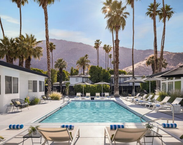A pool area with lounge chairs and umbrellas, surrounded by palm trees and mountains in the background.