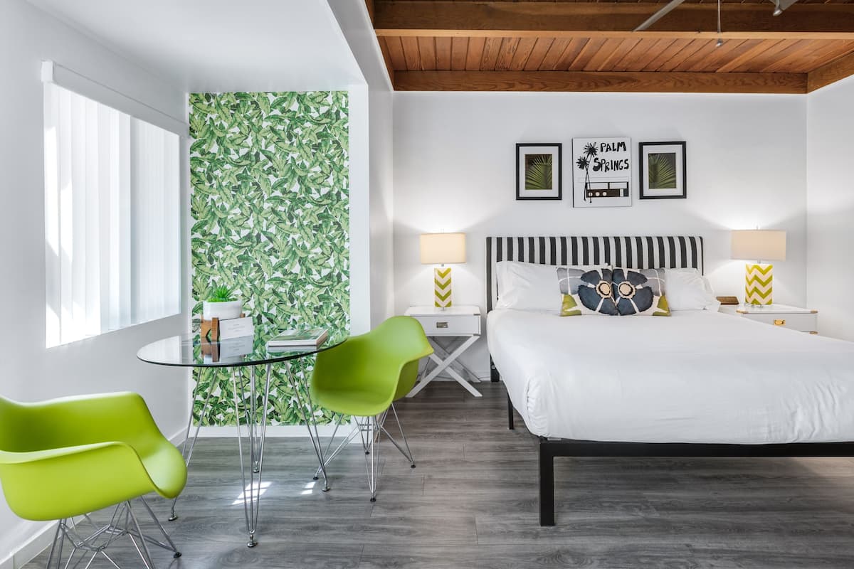 A modern bedroom with a bed, two bedside lamps, a green chair, a small table, and a wall with a leafy pattern.