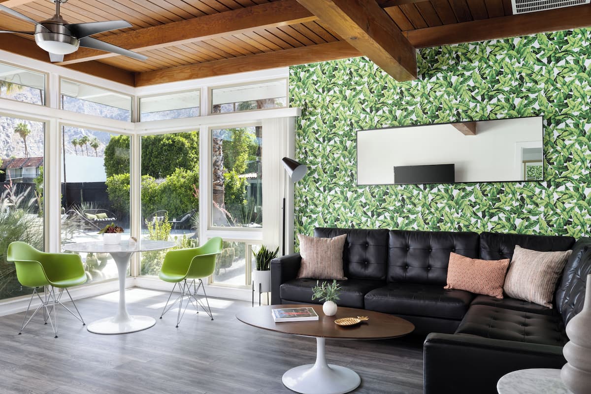 A modern living room with a black leather sofa, green chairs, a round coffee table, and a large window offering a view of greenery and mountains.