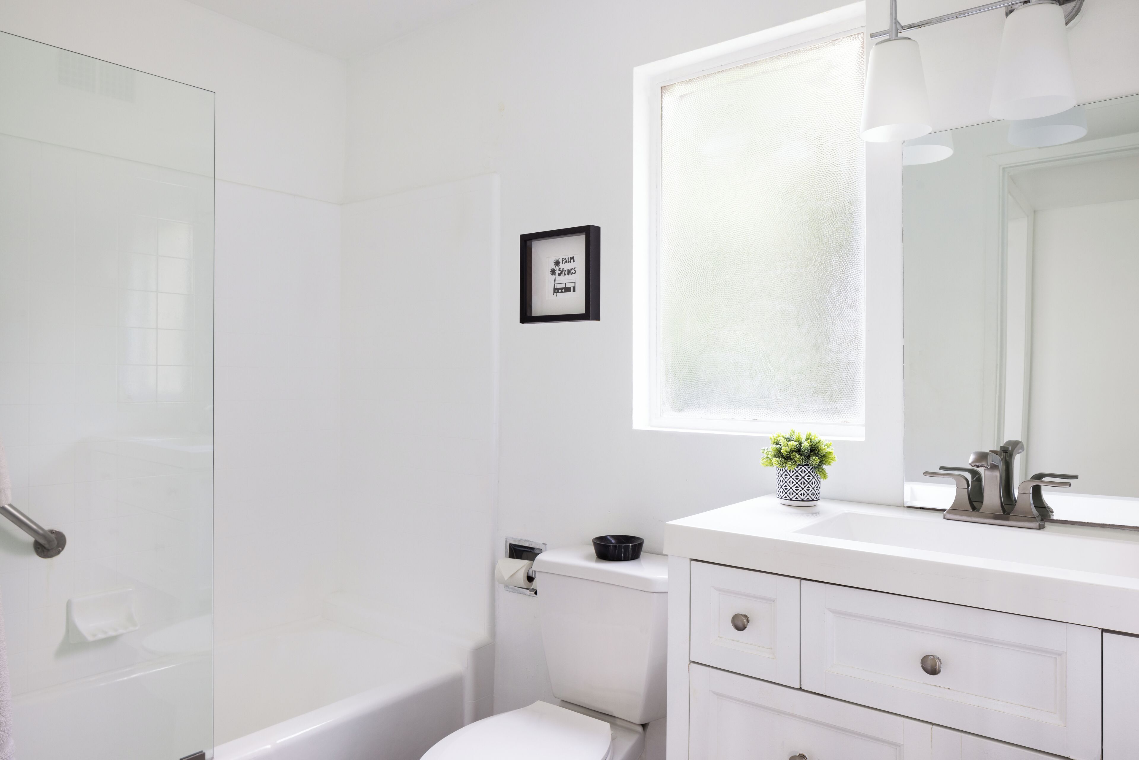 Et moderne badeværelse med brusekabine i glas, et hvidt toilet, en vask med spejl og en lille potteplante på bordet.