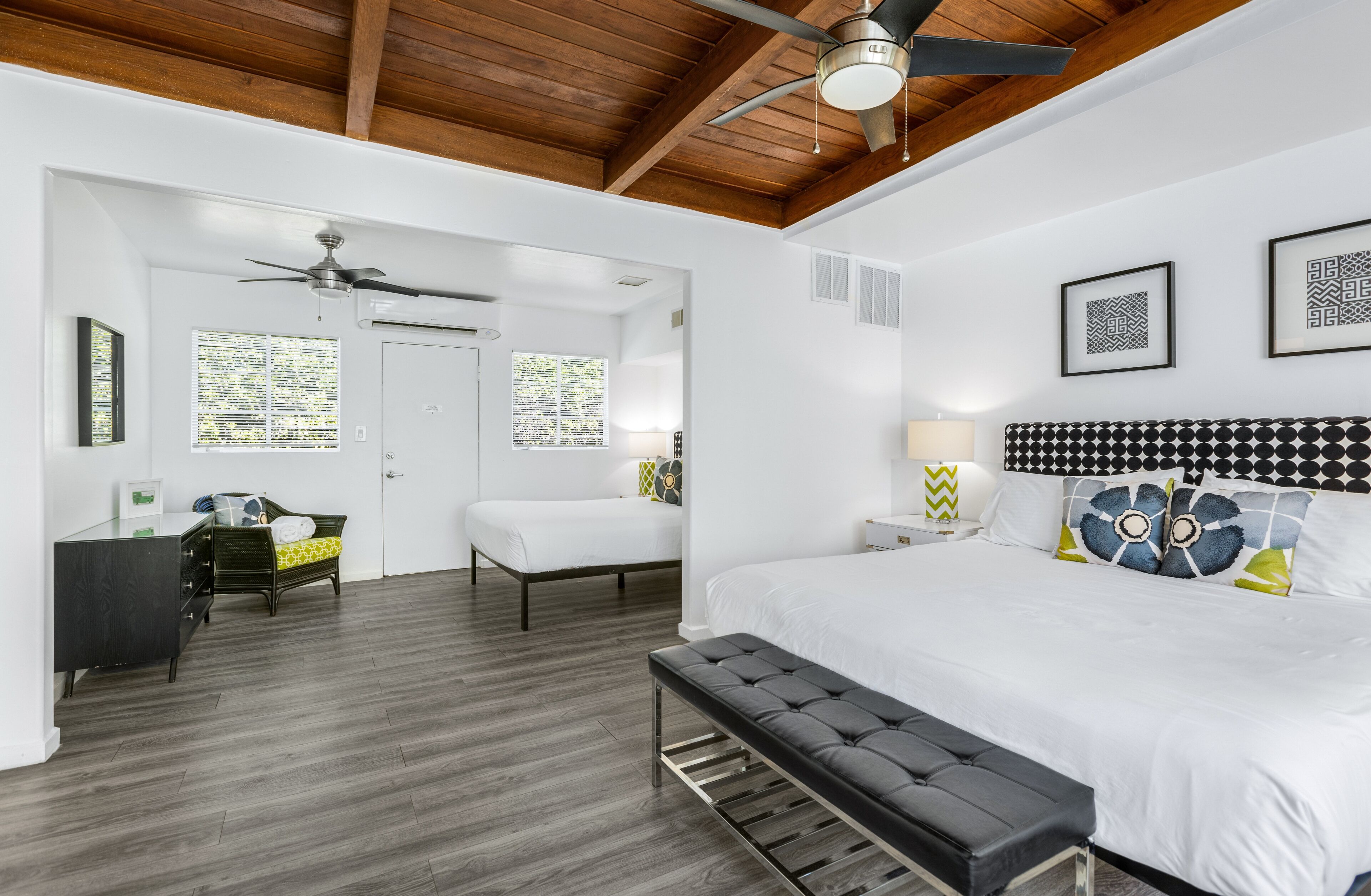 A bedroom with a large bed, a desk, a chair, and a bench. There is a ceiling fan and a window with blinds.