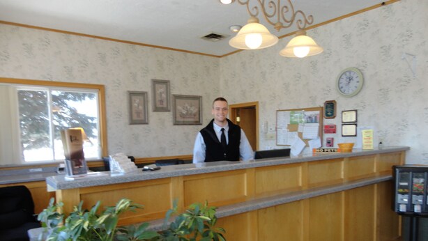 A reception desk with a wooden counter, framed pictures on the wall, a clock, and a bulletin board. - Bangor Inn & Suites (Bangor)