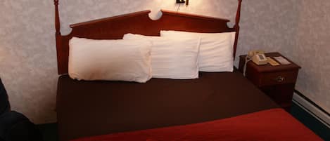 Cama bem arrumada com travesseiros brancos e um cobertor vermelho, uma luminária de cabeceira e um criado-mudo de madeira com telefone.