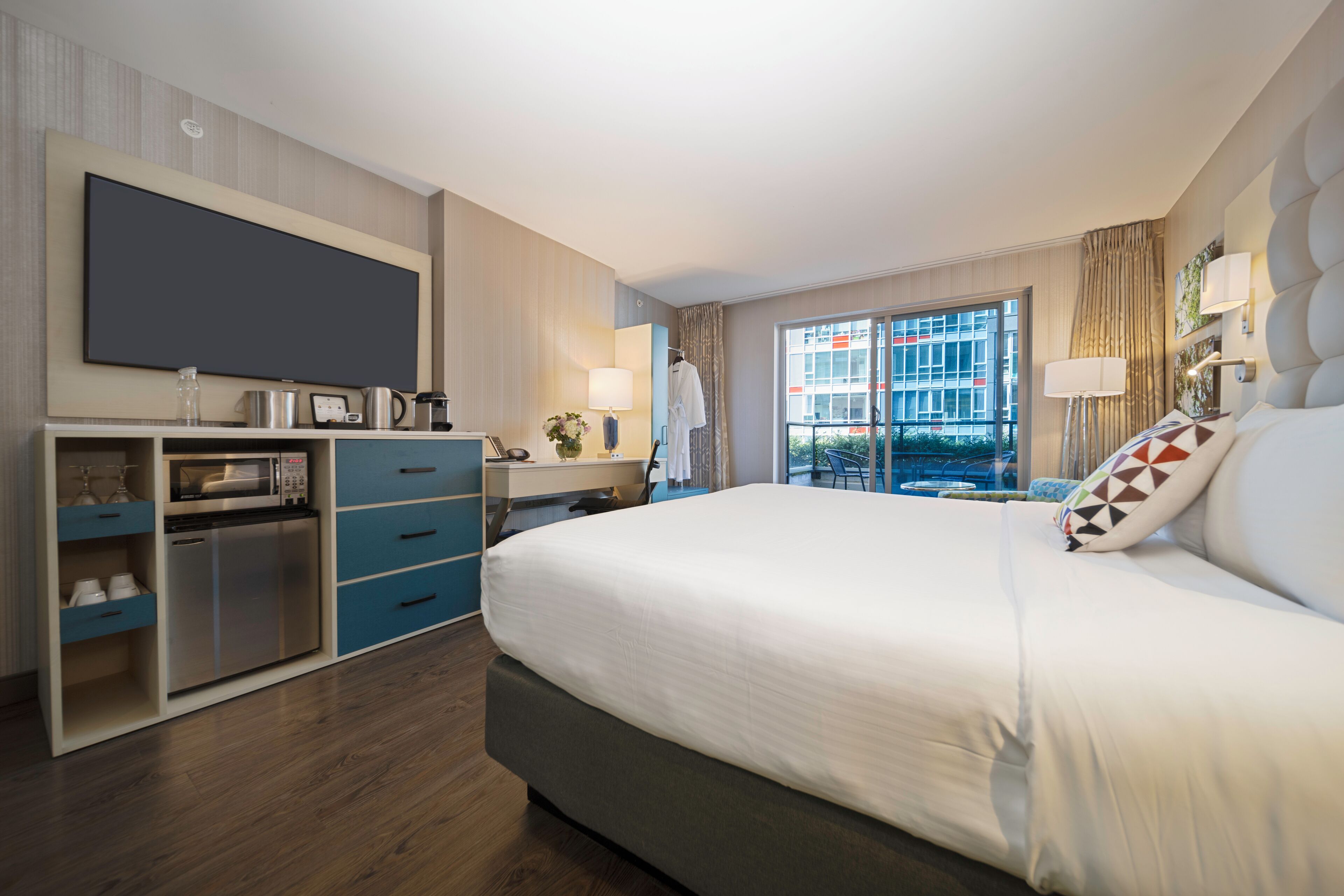 Ein modernes Hotelzimmer mit einem großen Bett, einem flachbildfernseher an der Wand, einem Schreibtisch mit Stuhl und Blick auf die Stadt durch das Fenster.