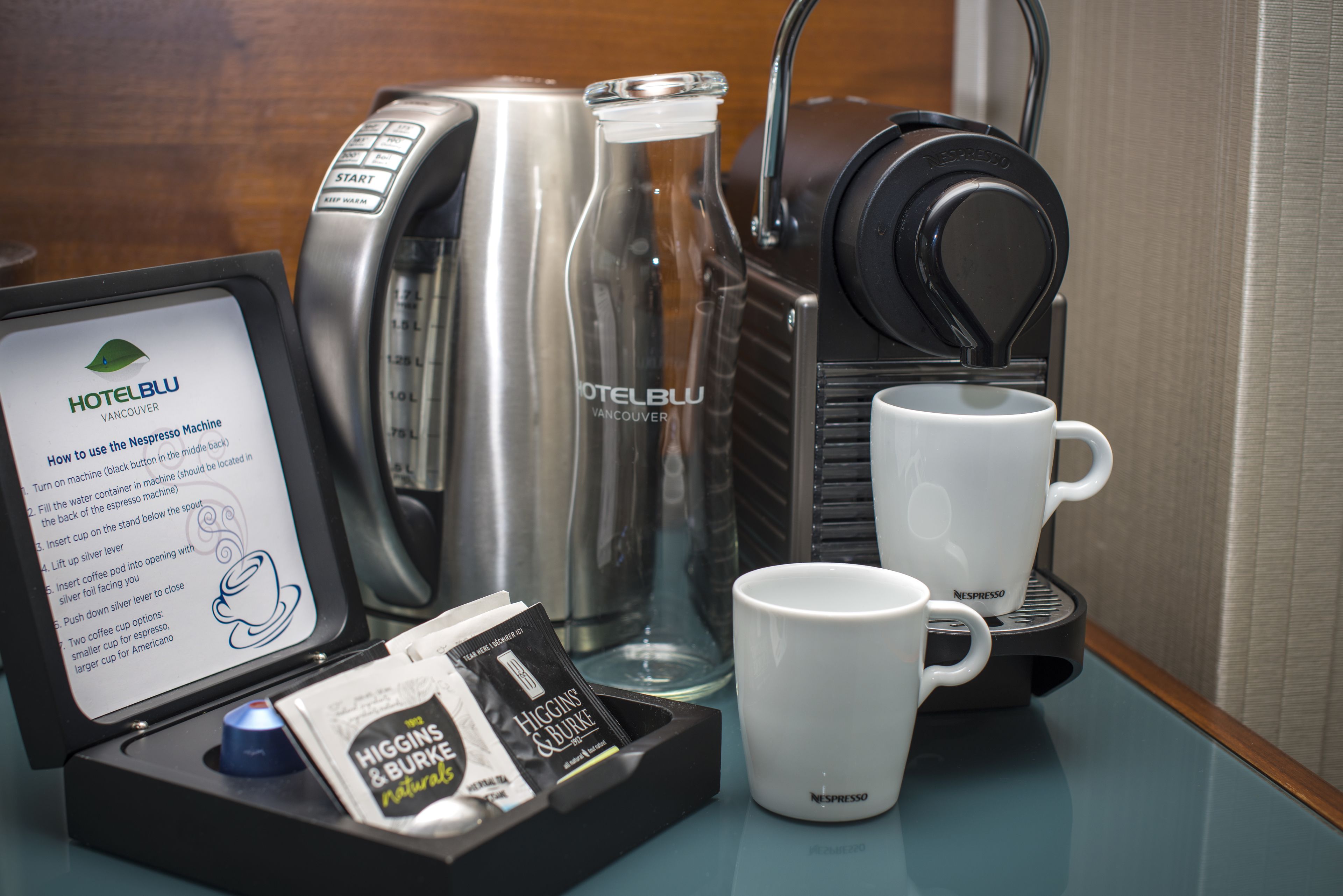 A coffee machine, two white mugs, a glass bottle, and a booklet on a table.
