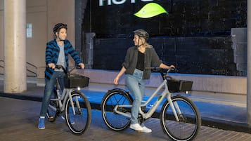 Due persone in bicicletta, con cestini, davanti a un edificio con un cartello che riporta la scritta 'HOTELBLO'.