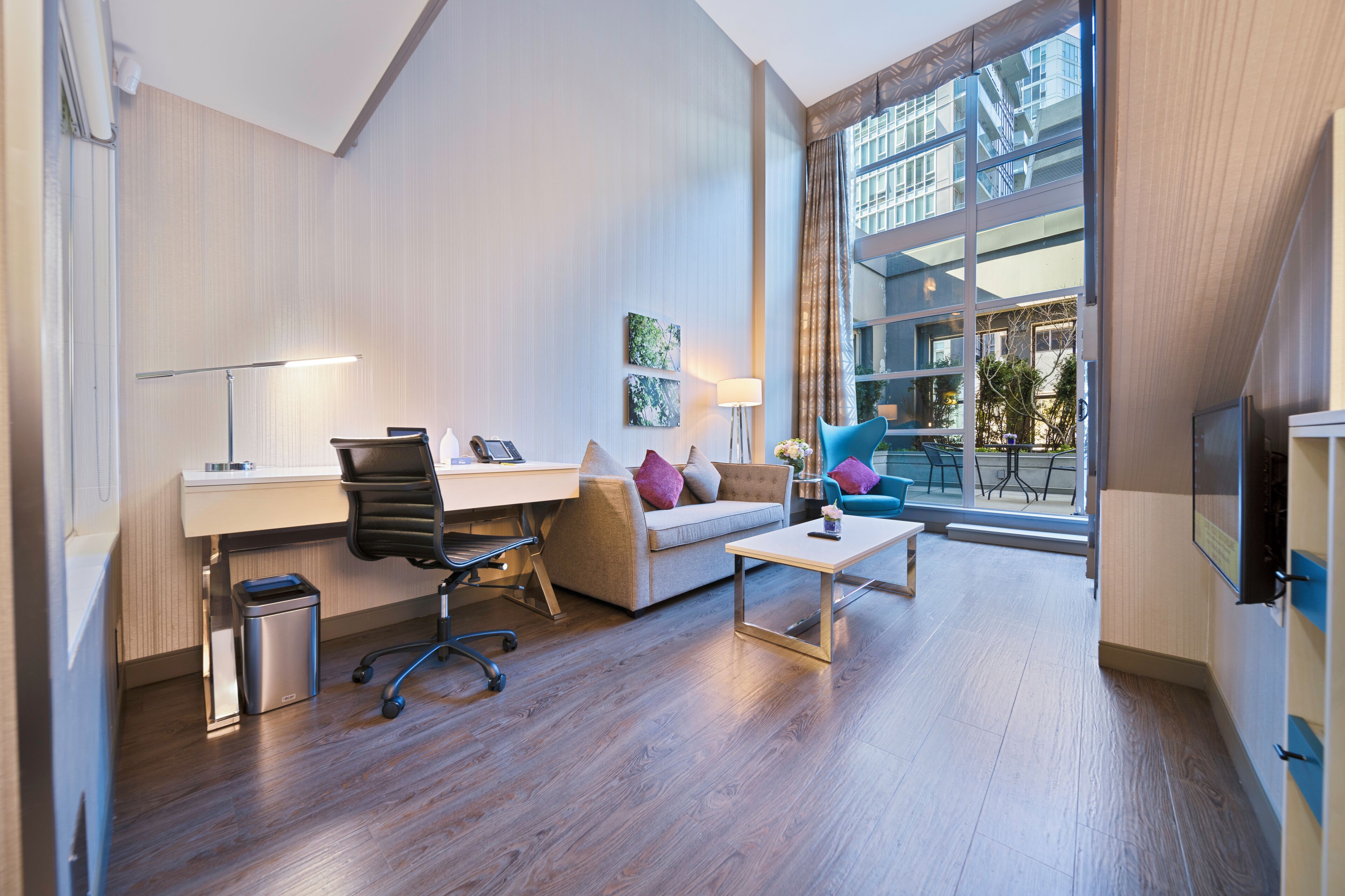 Una habitación de hotel moderna con un escritorio, una silla de oficina, un sofá y una pequeña mesa. Se observa un cesto de basura, una lámpara y un televisor. La habitación tiene ventanas amplias con vistas a un edificio.