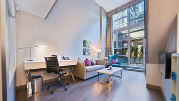Una habitación de hotel moderna con un escritorio, una silla de oficina, un sofá y una pequeña mesa. Se observa un cesto de basura, una lámpara y un televisor. La habitación tiene ventanas amplias con vistas a un edificio.