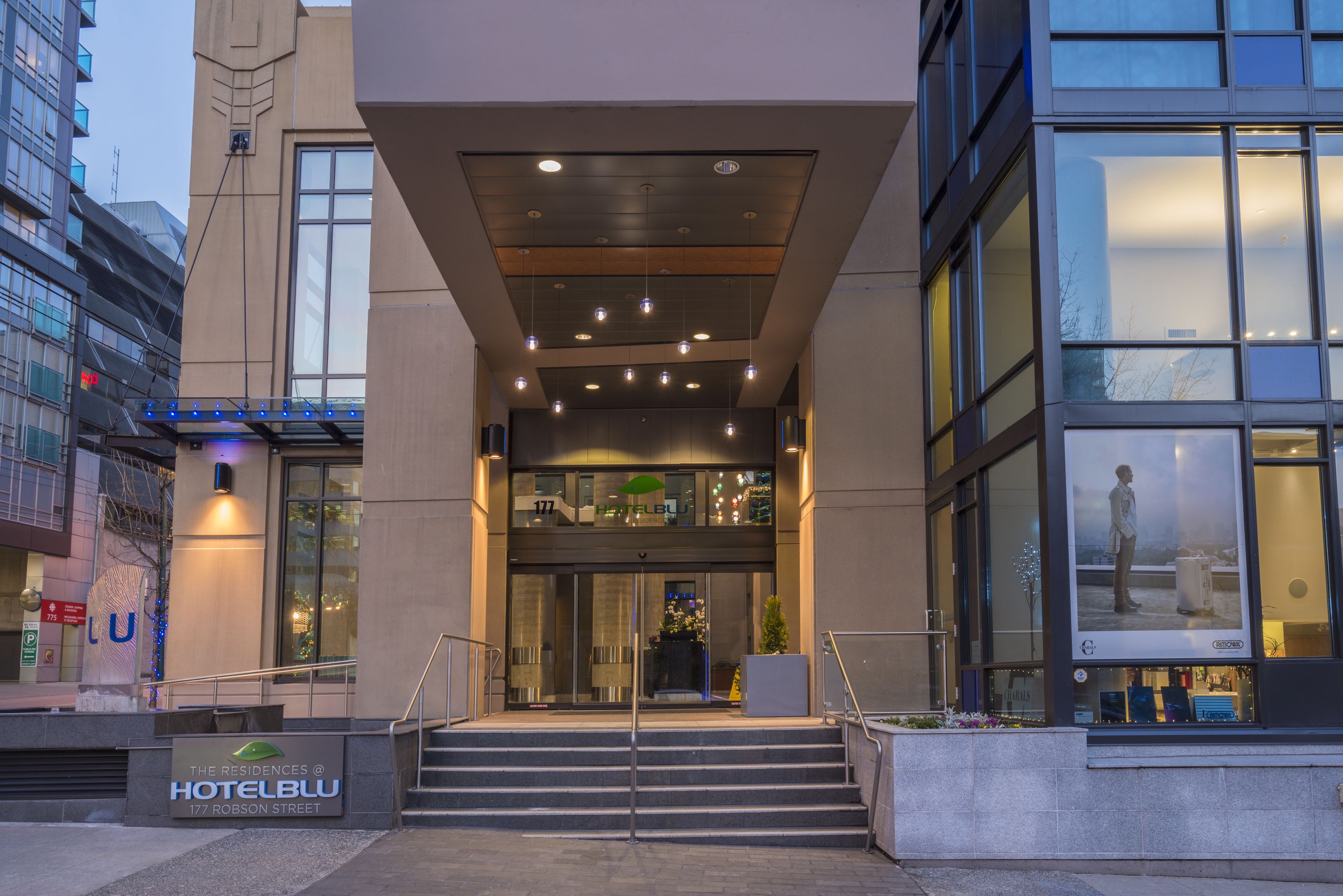 A modern hotel entrance with a staircase leading to the lobby, large windows, and a sign indicating the hotel name and address.