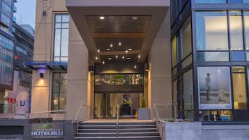 A modern hotel entrance with a staircase leading to the lobby, large windows, and a sign indicating the hotel name and address.