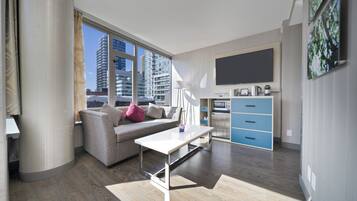 A modern living room with a sofa, a coffee table, and a TV. There is a kitchen area with cabinets and a microwave.