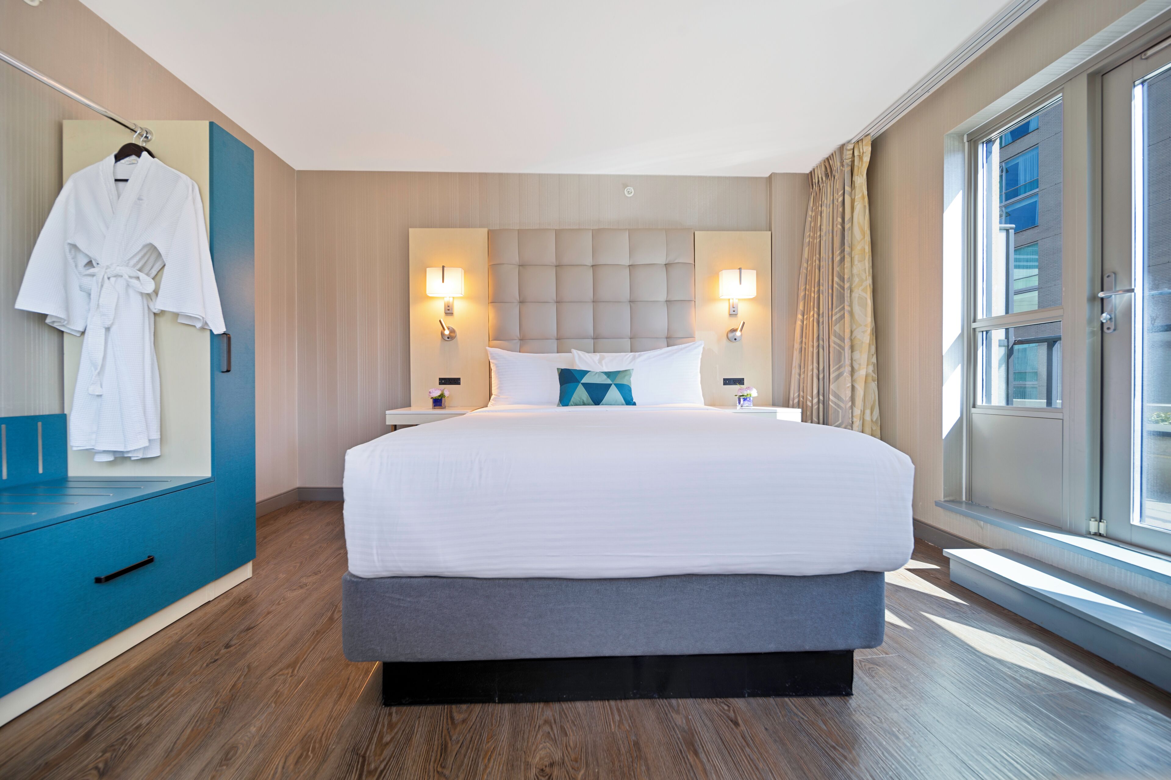 A modern hotel room with a large bed, two bedside lamps, a robe hanging on the wall, and a view of the city through the window.