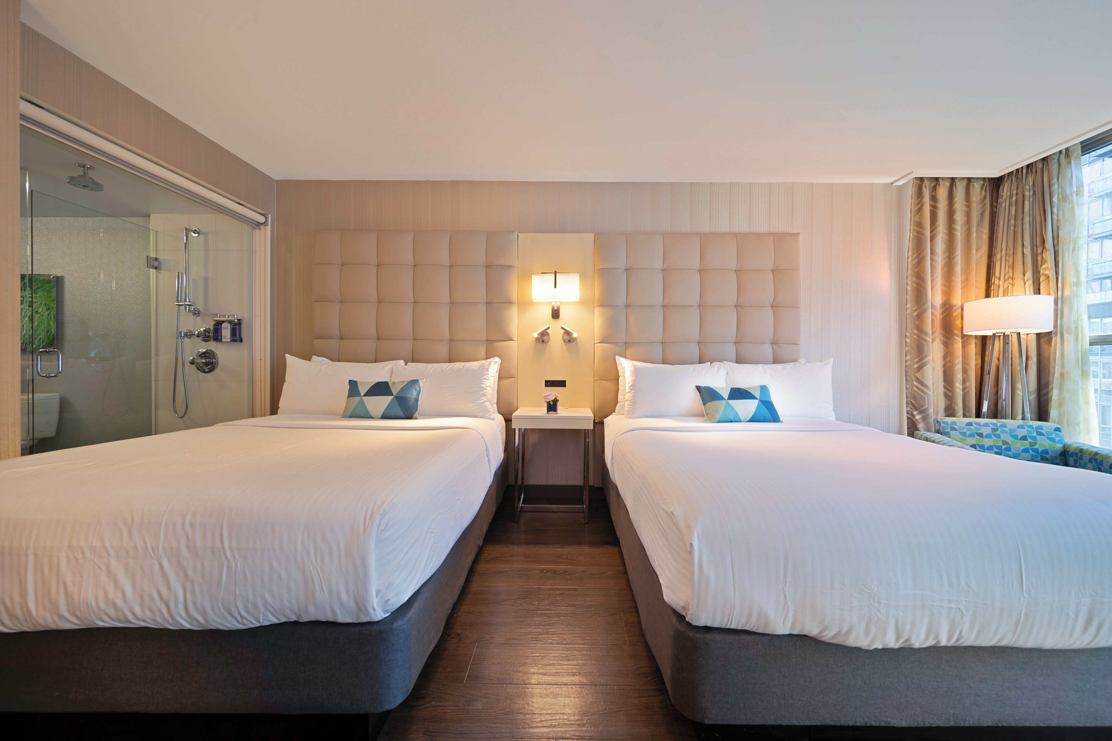A hotel room with two beds, a glass shower enclosure, a wall-mounted light fixture, and a view of the city through large windows.