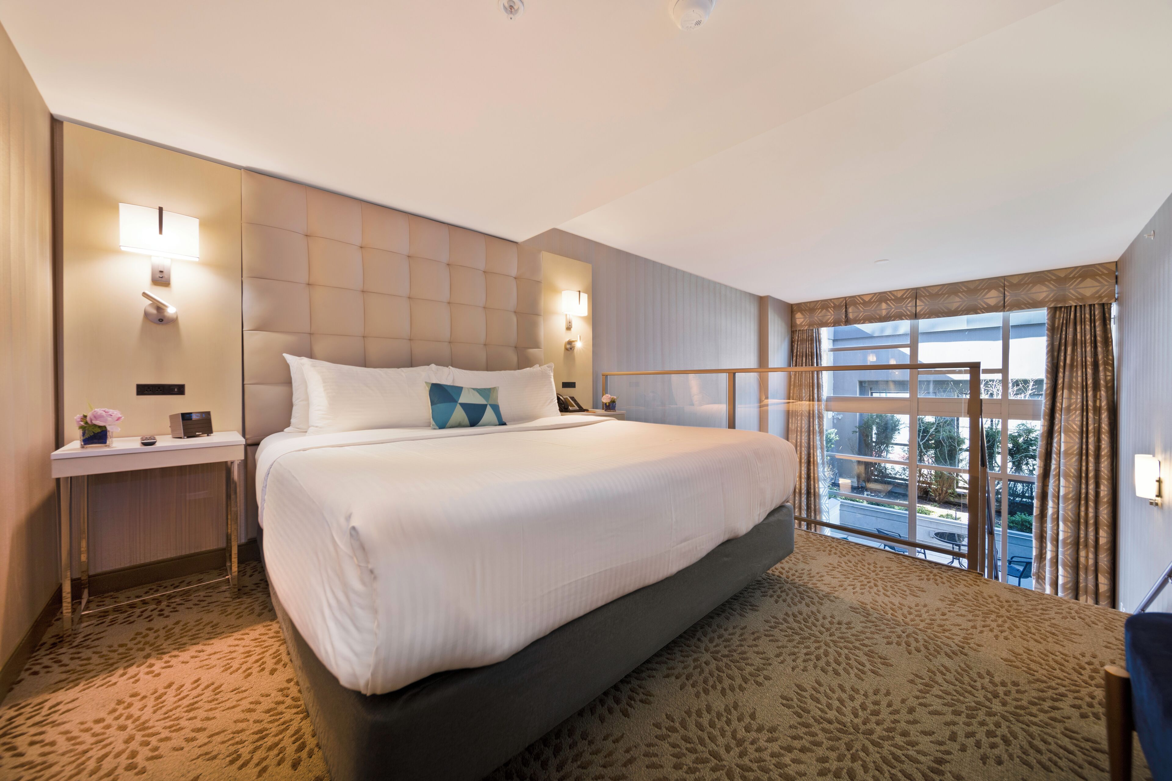 Habitación de hotel con una cama grande, una mesita de noche con lámpara, una ventana con vista al exterior y piso alfombrado.