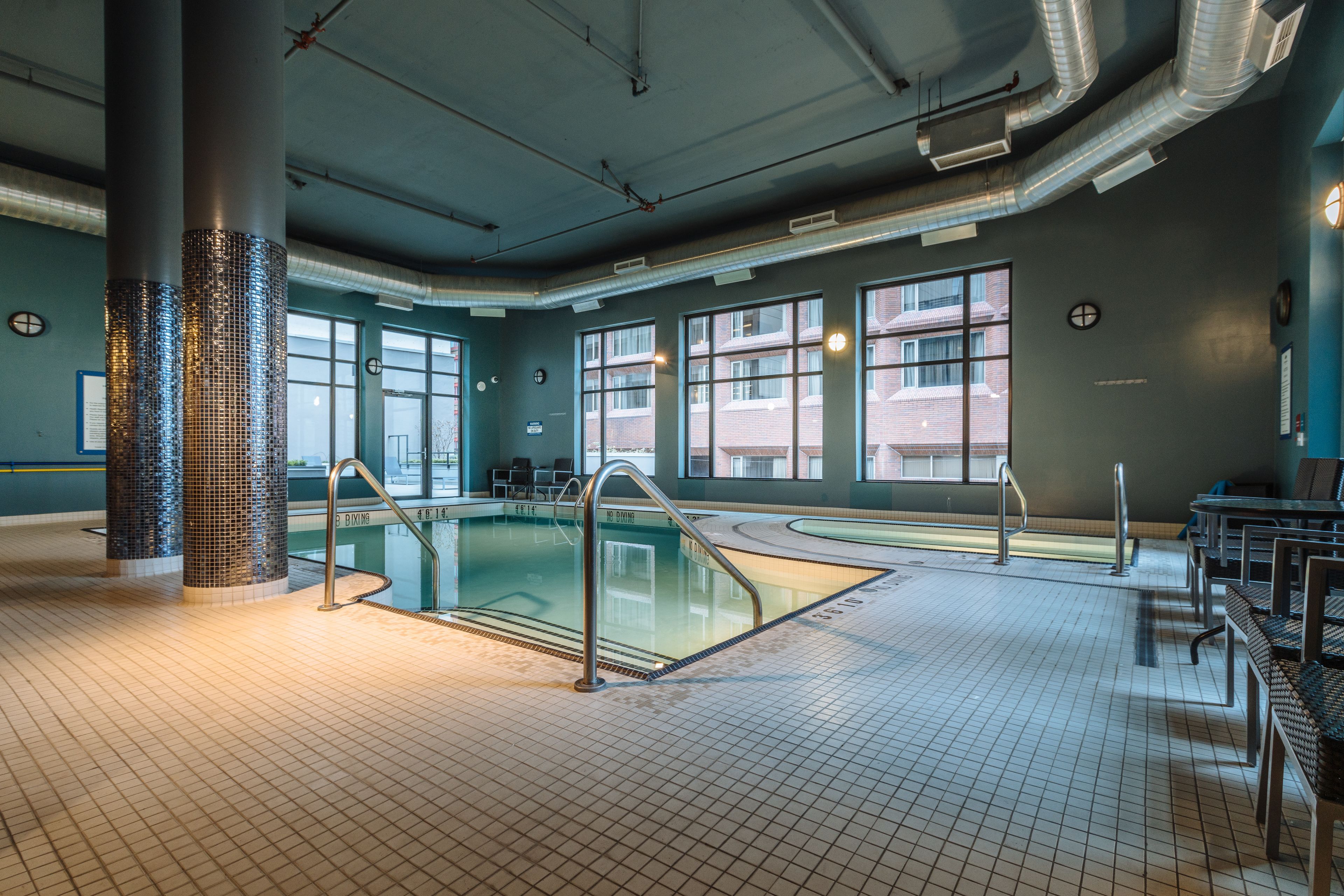 A swimming pool with tiled flooring and metal railings, large windows, and a modern ceiling with visible ductwork.