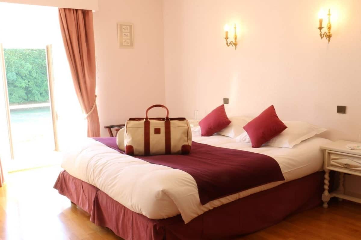 Un lit bien fait, recouvert d’une couverture rouge et blanche, une grande valise posée sur le lit et une table de chevet avec une lampe.