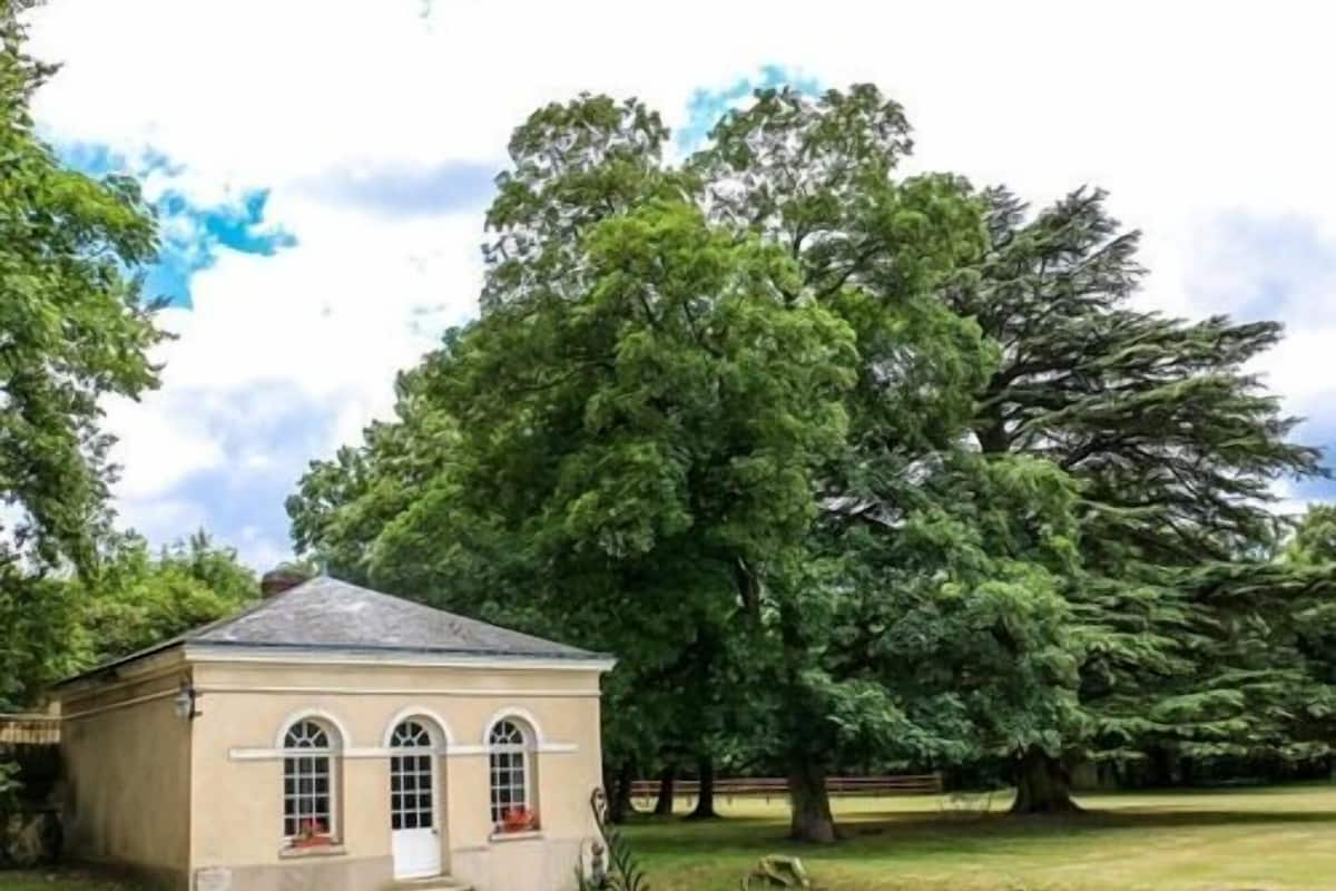 Un petit bâtiment d’un seul étage, à la façade beige et aux fenêtres en arc de cercle, entouré d’une végétation luxuriante et d’arbres.