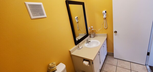 Un baño con una pared amarilla, un lavabo blanco, un espejo, un secador de pelo y un inodoro.