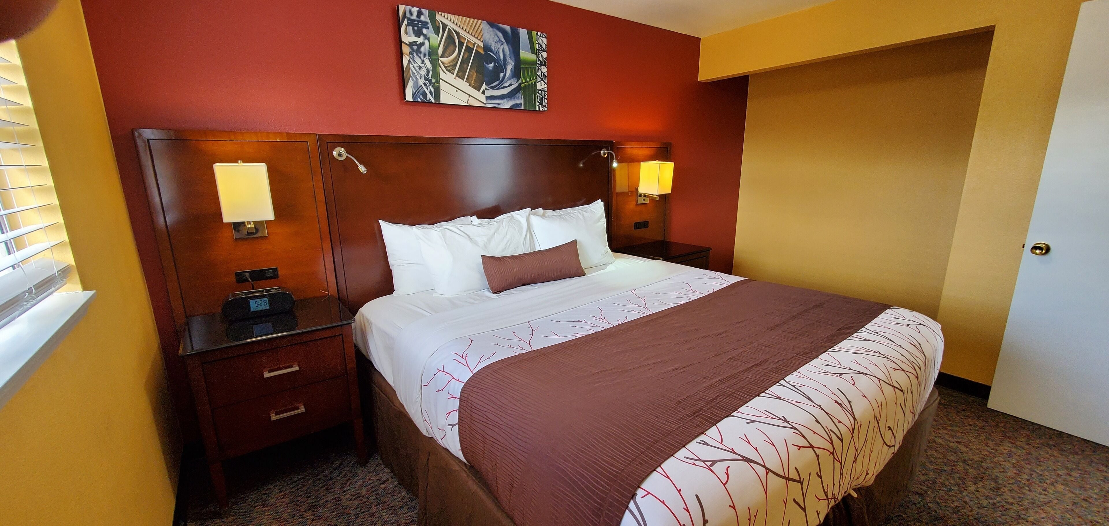 Habitación de hotel con una cama grande, dos lámparas de noche, una mesita de noche, un cuadro en la pared y una puerta.