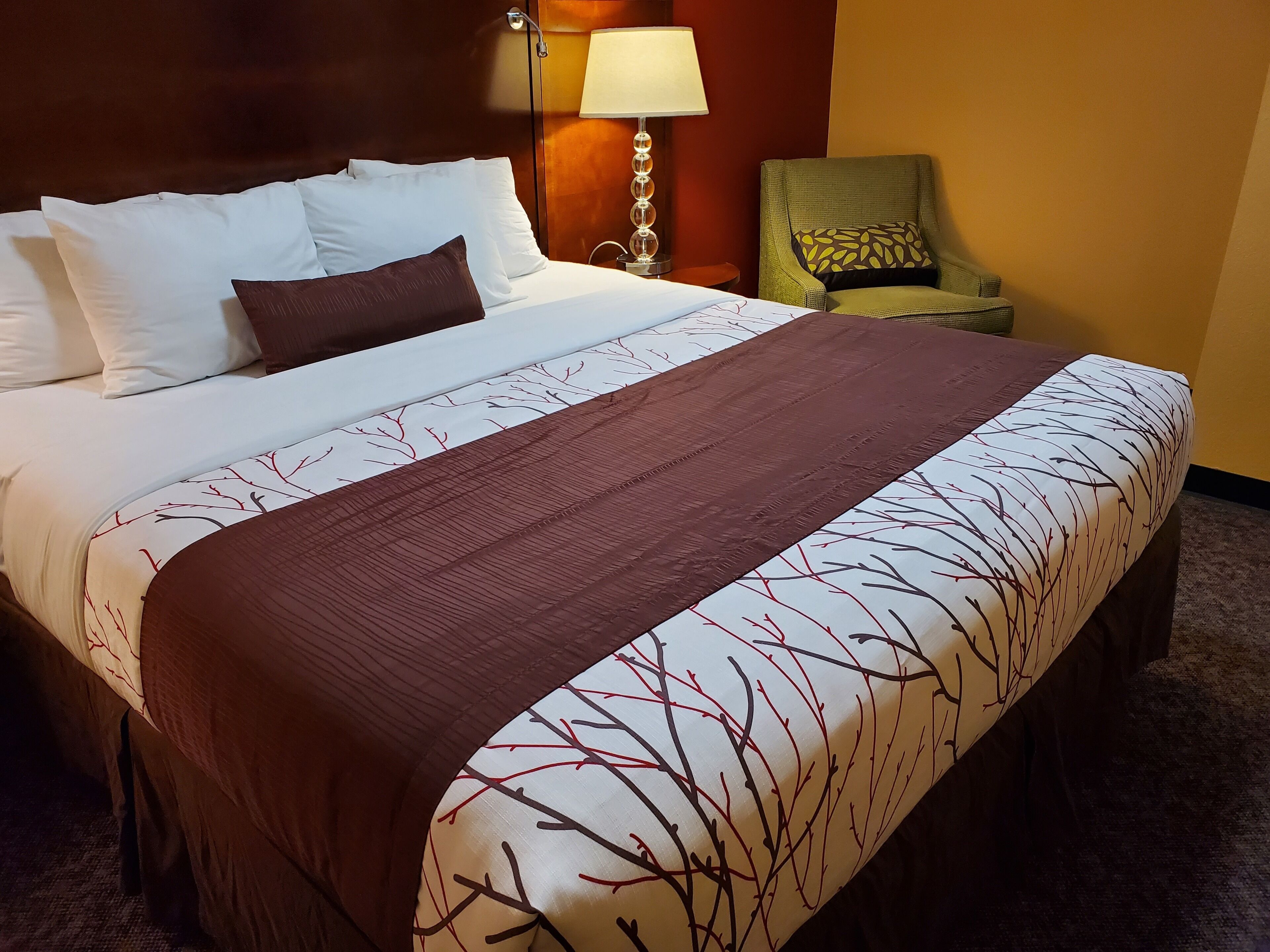 A neatly made bed with white and brown bedding, a green armchair with a patterned cushion, and a lamp on a bedside table.