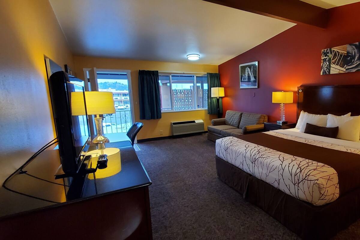 A hotel room with a large bed, a desk with a lamp, a chair, a window with curtains, and a painting on the wall.