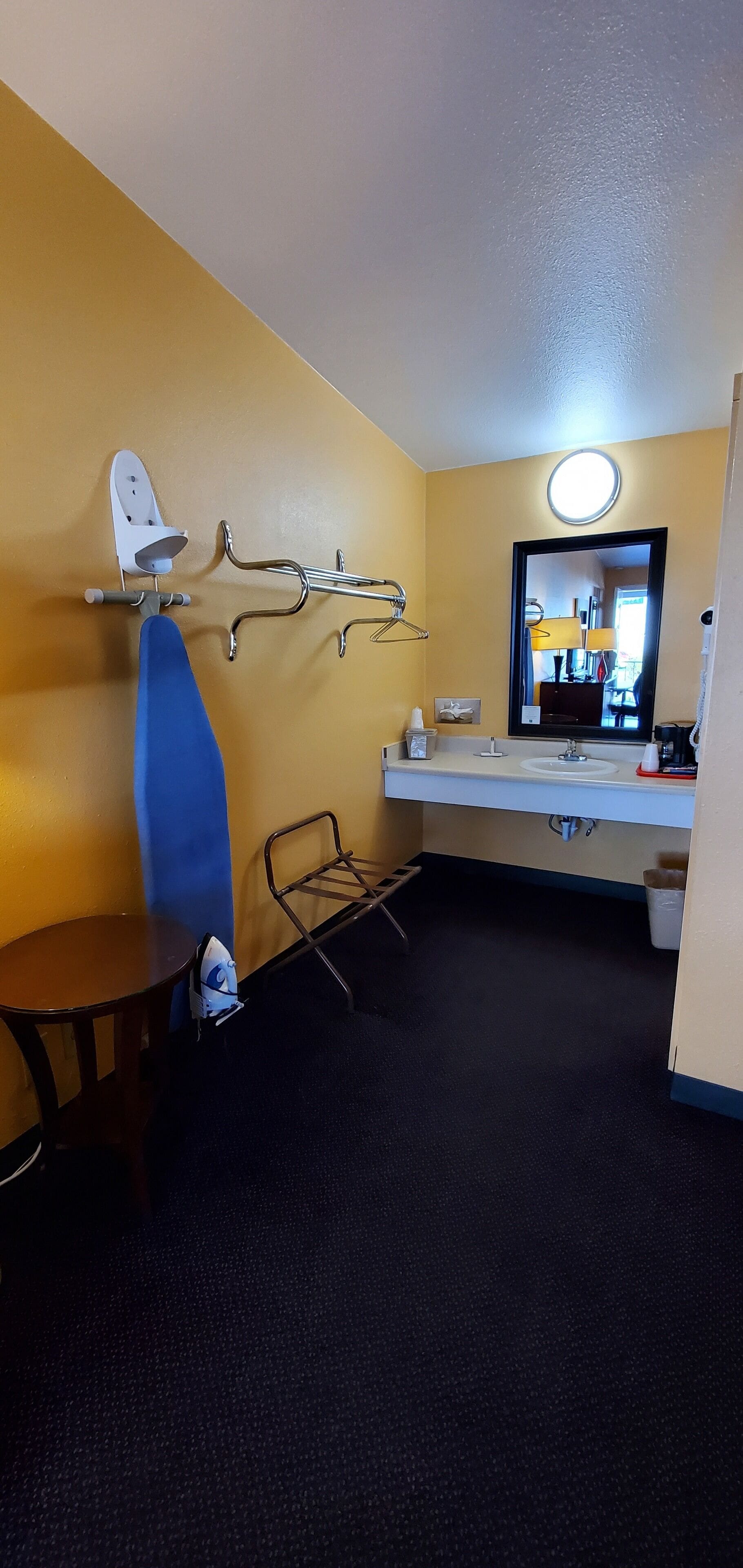 A compact hotel room with a single bed, a small table, a chair, a desk, a mirror, and a bathroom with a sink and toilet.