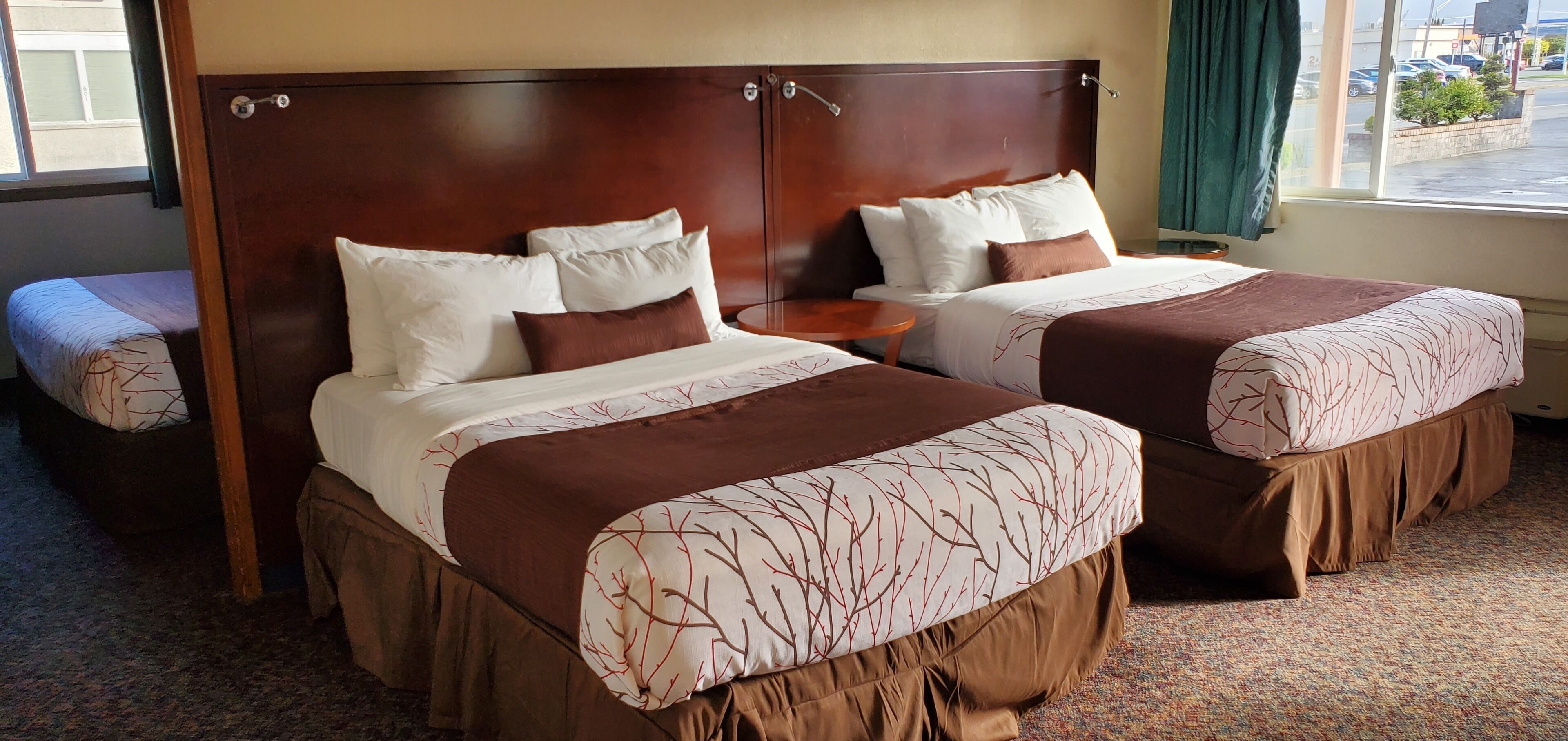 A hotel room with two beds, a small round table, and a view of the street through a window.