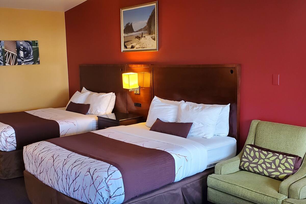 A hotel room with two beds, a green armchair, and a painting on the wall.
