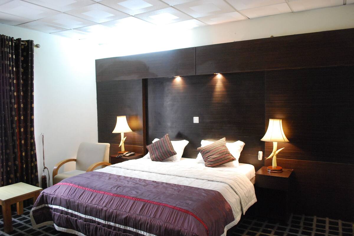A hotel room with a large bed, two bedside tables with lamps, a chair, and a curtain.