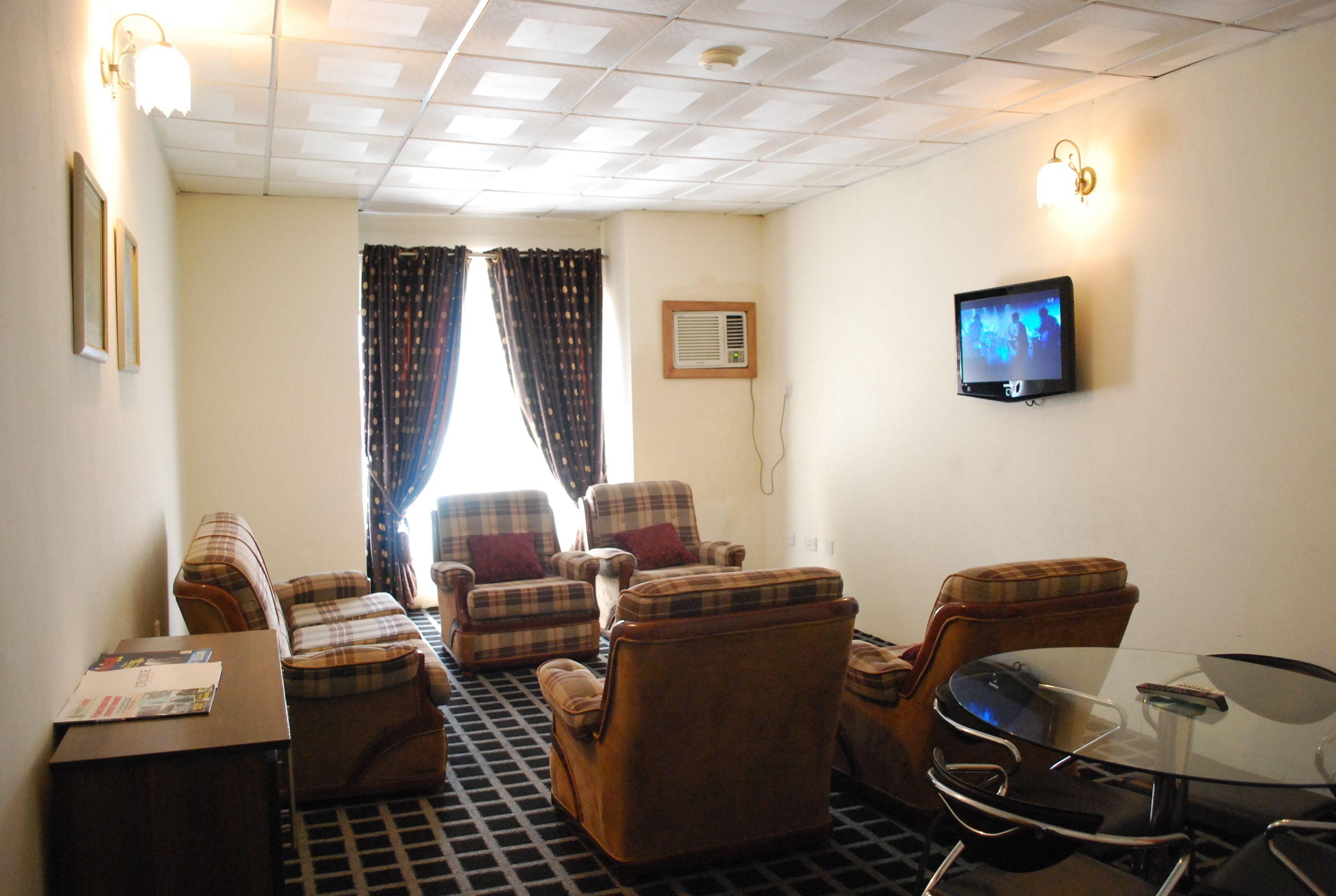 A hotel room with a checkered floor, plaid-patterned chairs, a small table, a television on the wall, and a window with curtains.