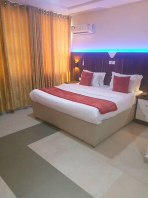 A hotel room with a large bed, a nightstand, an air conditioning unit, and a curtain. - Citilodge Hotel Lagos (Lekki)