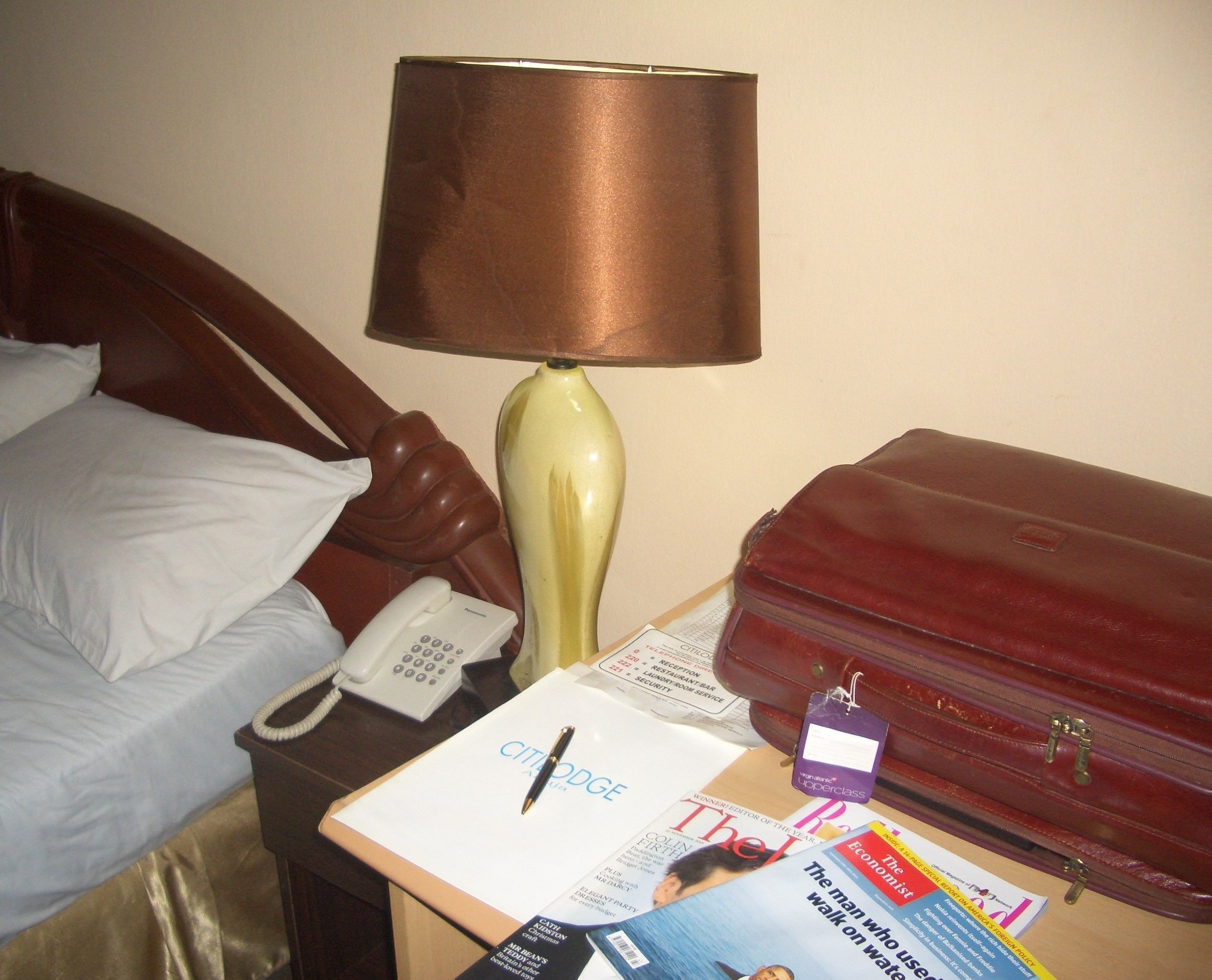 A hotel room with a bed, a lamp, a telephone, a magazine, and a suitcase.