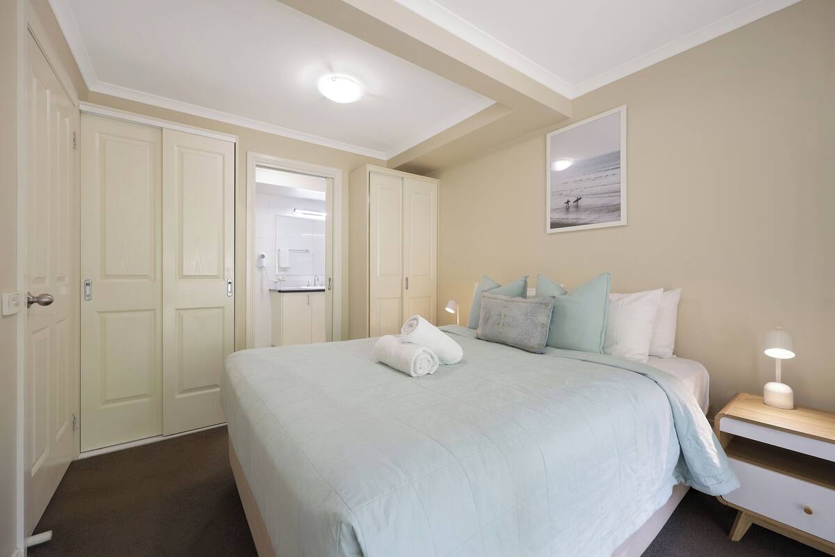 A bedroom with a bed, a nightstand, a wall-mounted lamp, and a door leading to another room.