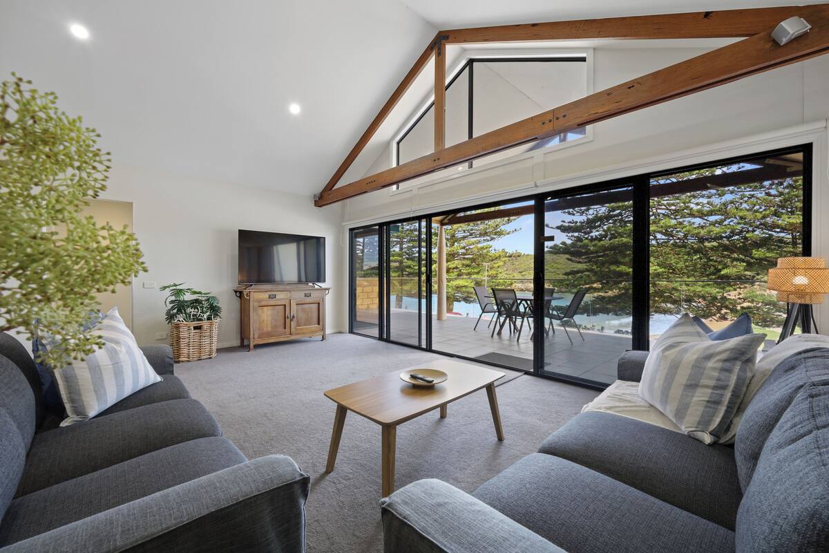 A modern living room with a sofa, a coffee table, and a television. There is a large window with a view of trees and a patio area.
