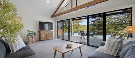 A modern living room with a sofa, a coffee table, and a television. There is a large window with a view of trees and a patio area.