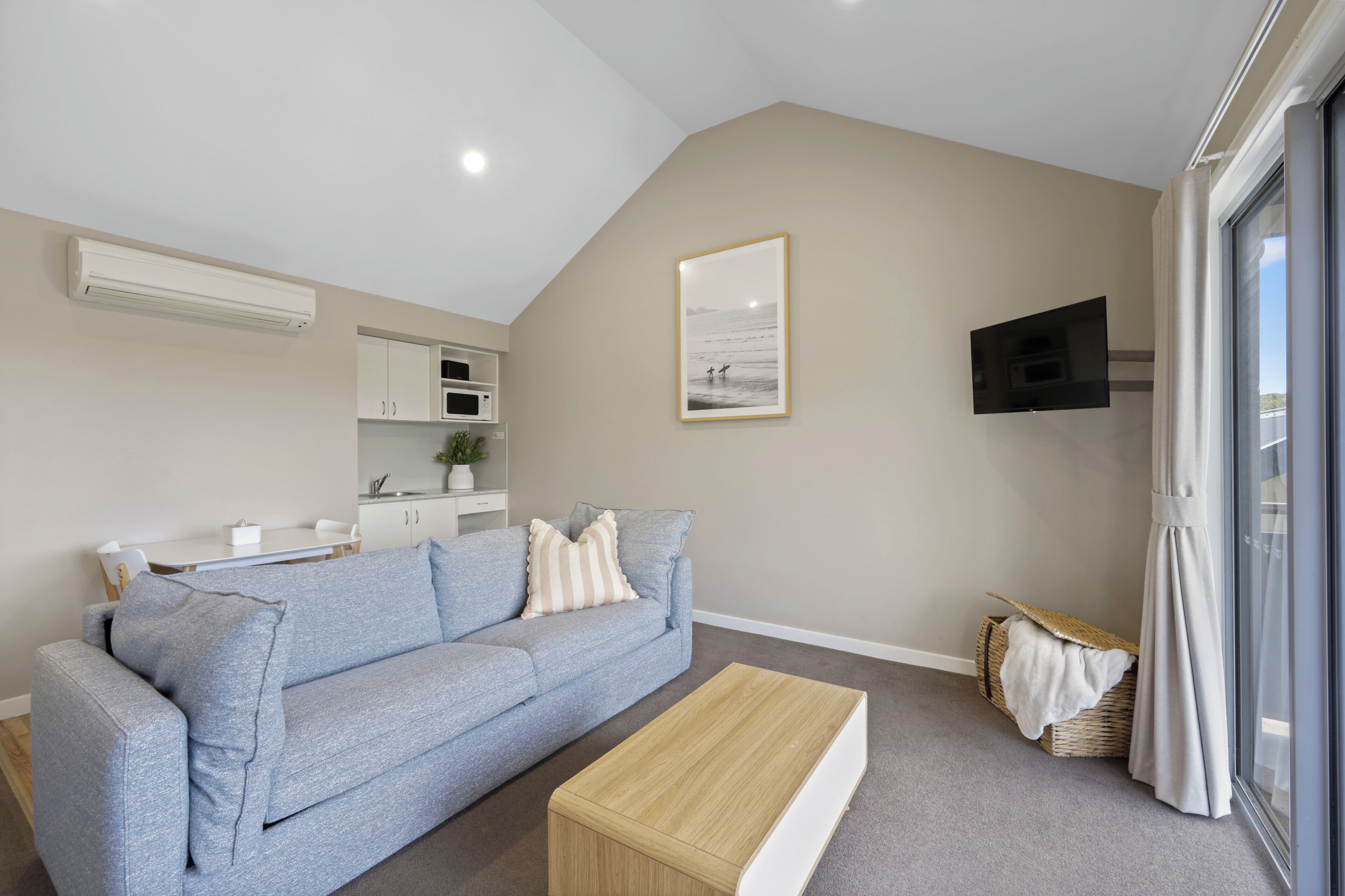 A modern living room with a grey sofa, a wooden coffee table, a kitchen area with white cabinets, a flat-screen TV mounted on the wall, and a large window with curtains.