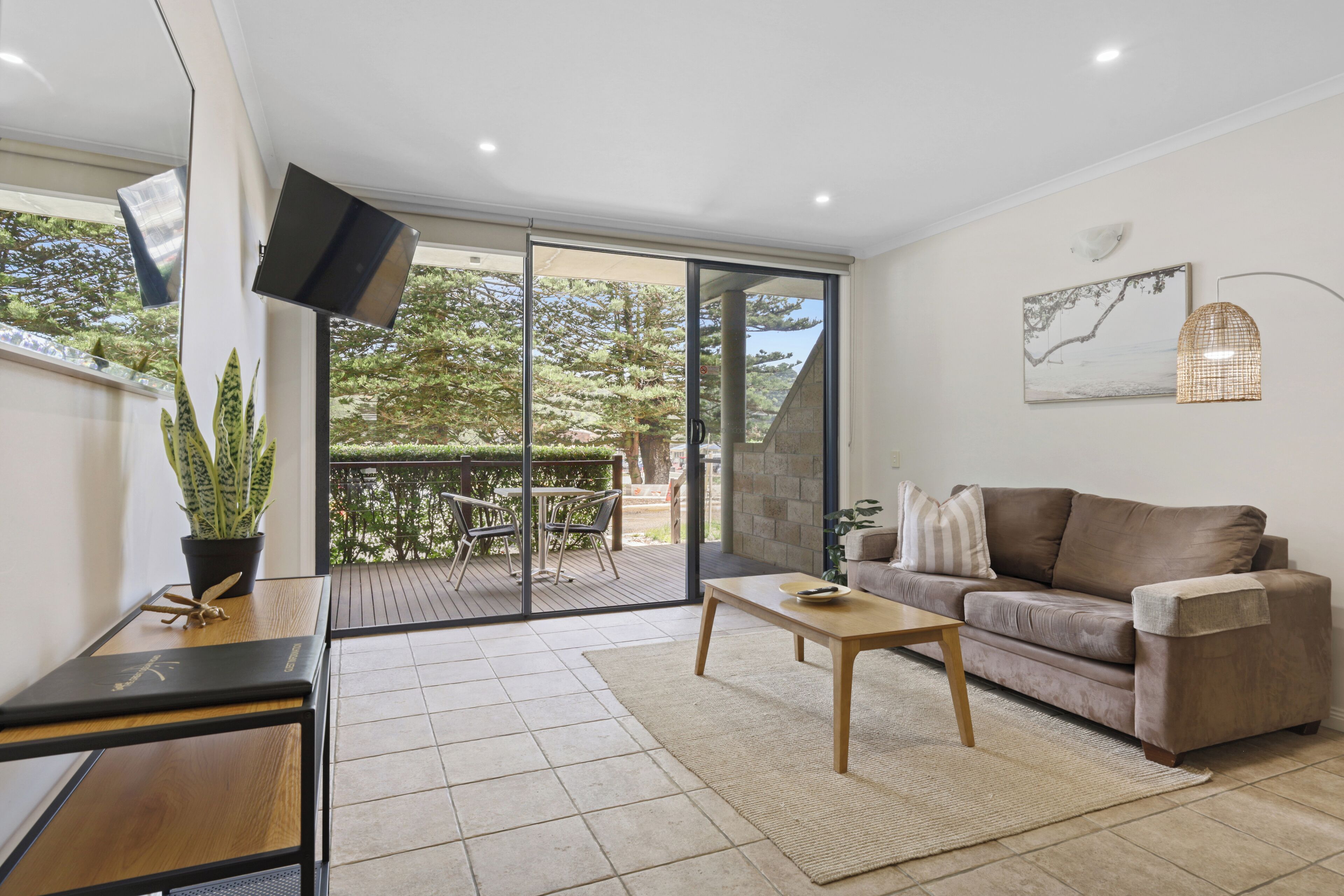 A modern living room with a sliding glass door leading to a balcony, a sofa, a coffee table, and a flat-screen TV mounted on the wall.