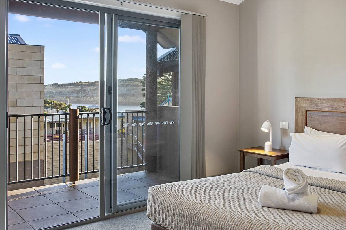 A hotel room with a bed, a nightstand, a lamp, and a view of a balcony with a glass door.