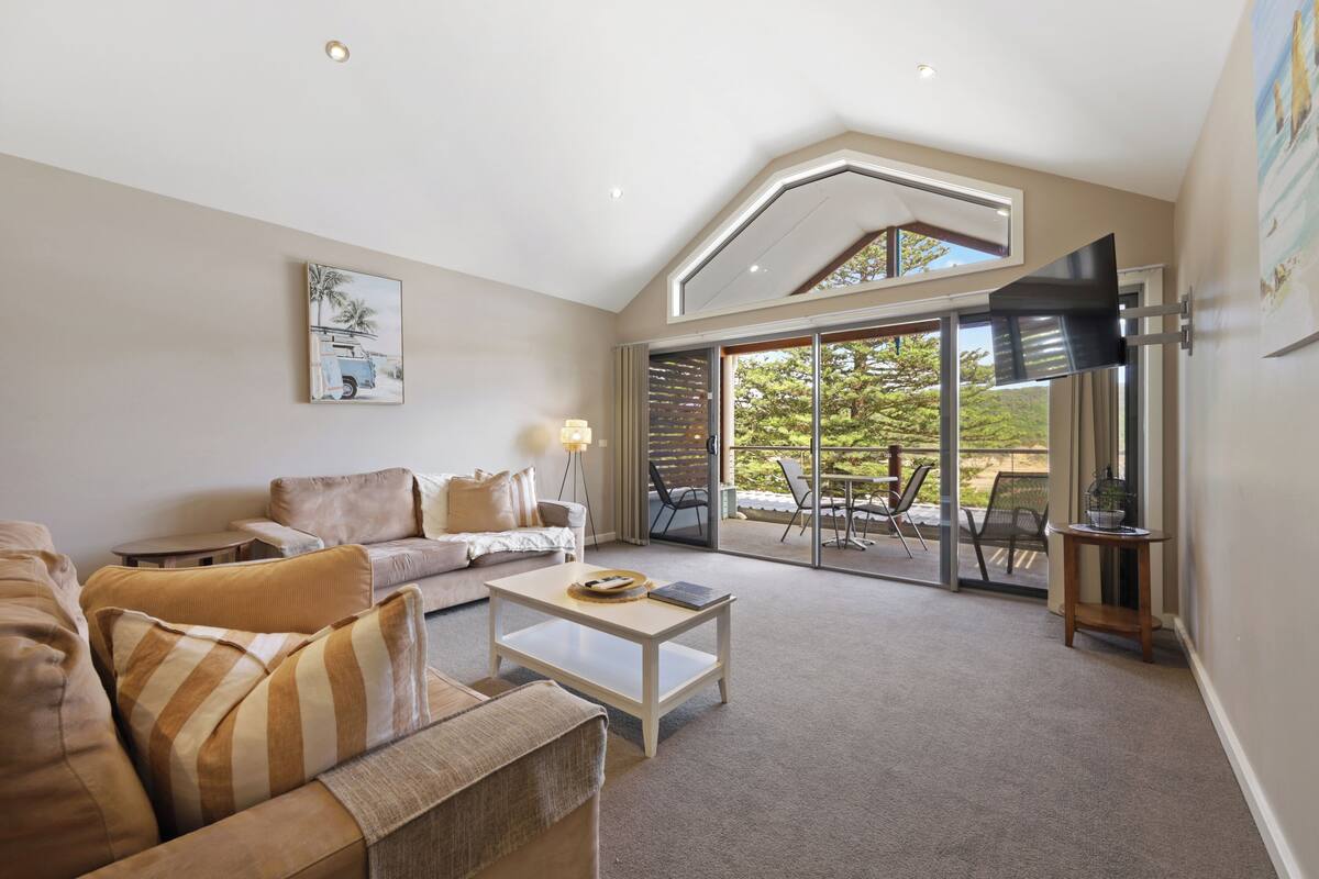 A spacious living room with a large sofa, a coffee table, and a television mounted on the wall.