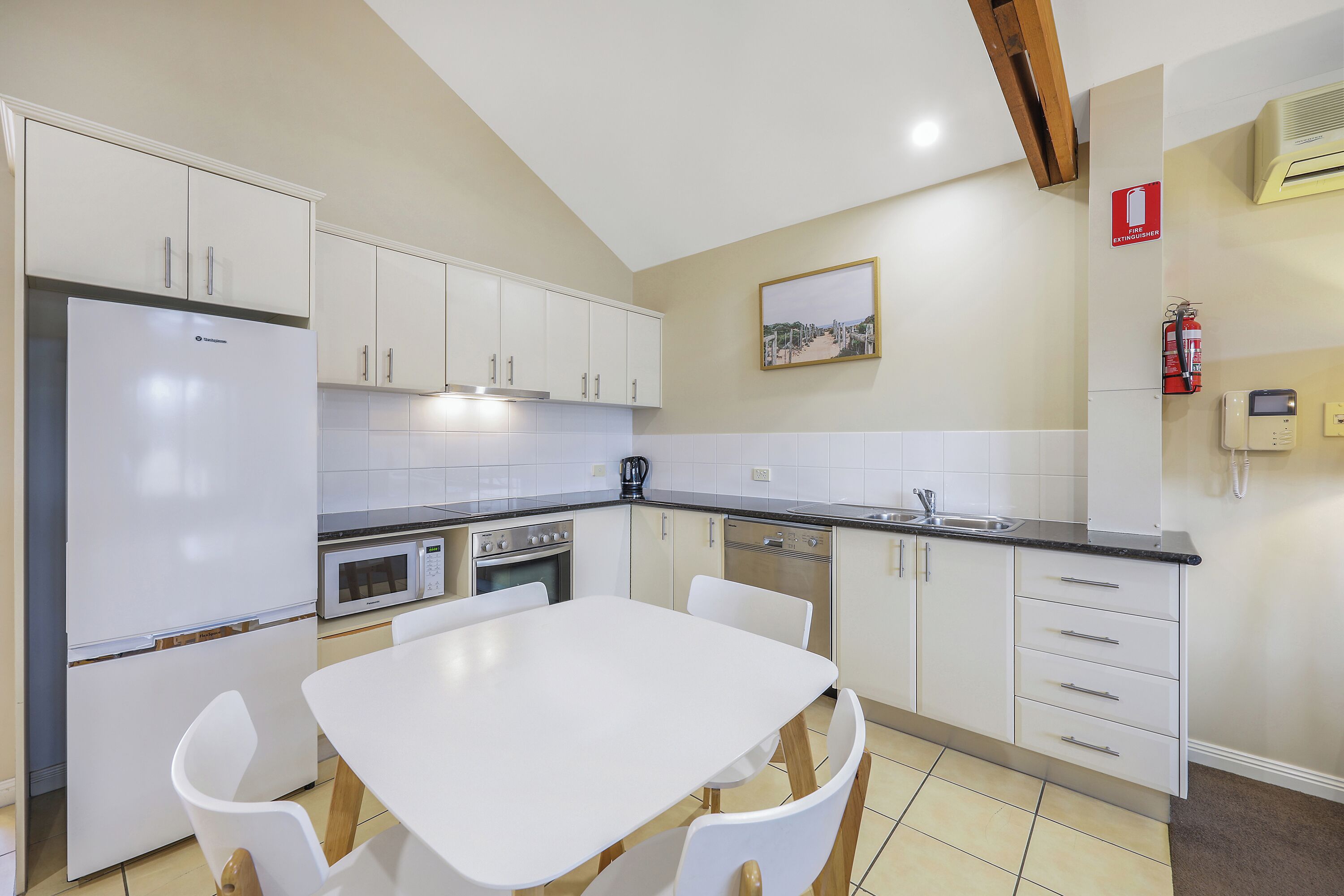 A modern kitchen with white cabinets, a stainless steel refrigerator, a microwave, an oven, and a dishwasher. There is a white dining table with four chairs.
