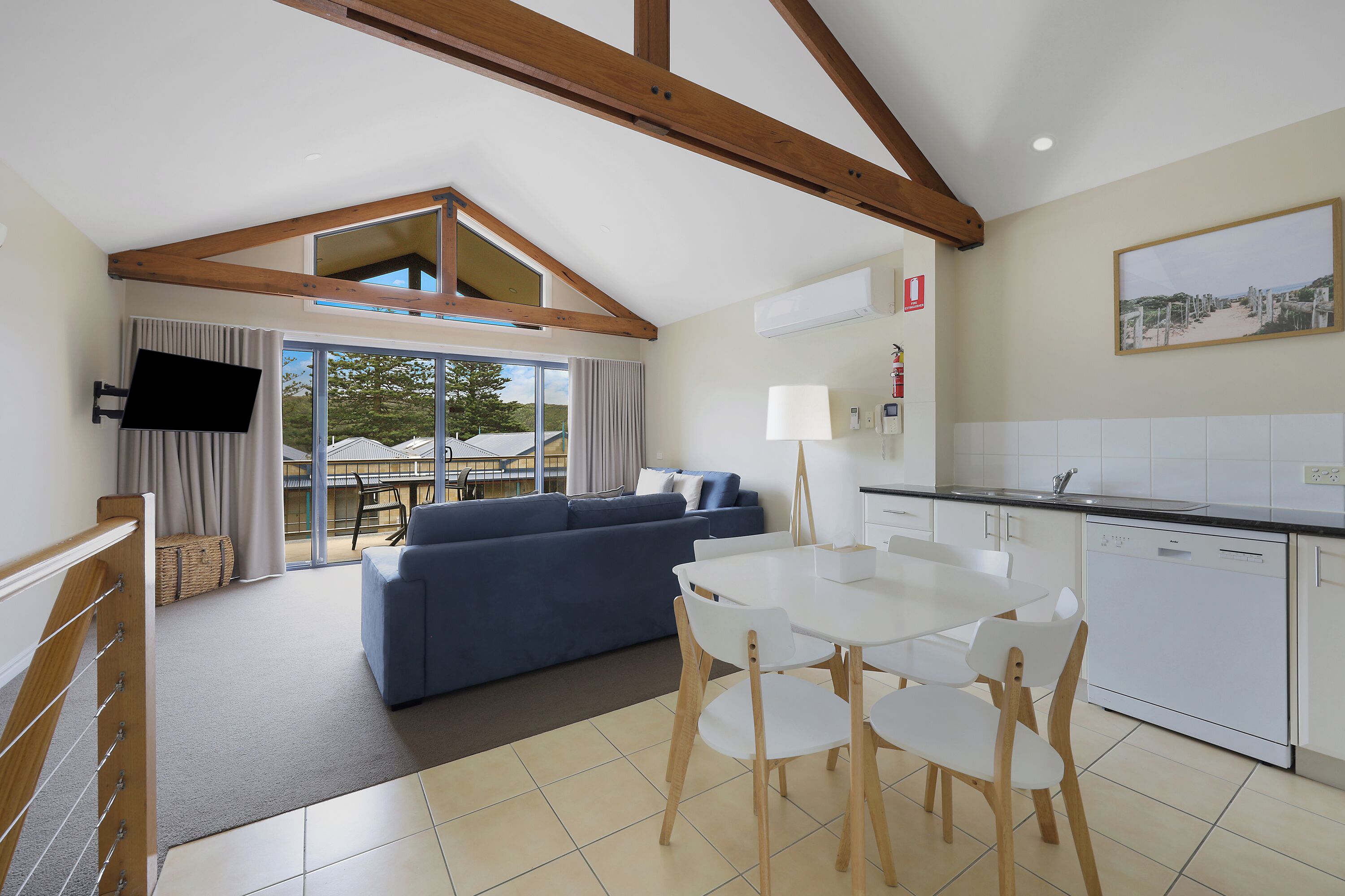 A modern living room with a blue sofa, a white dining table with chairs, and a kitchen area with white cabinets and a dishwasher.