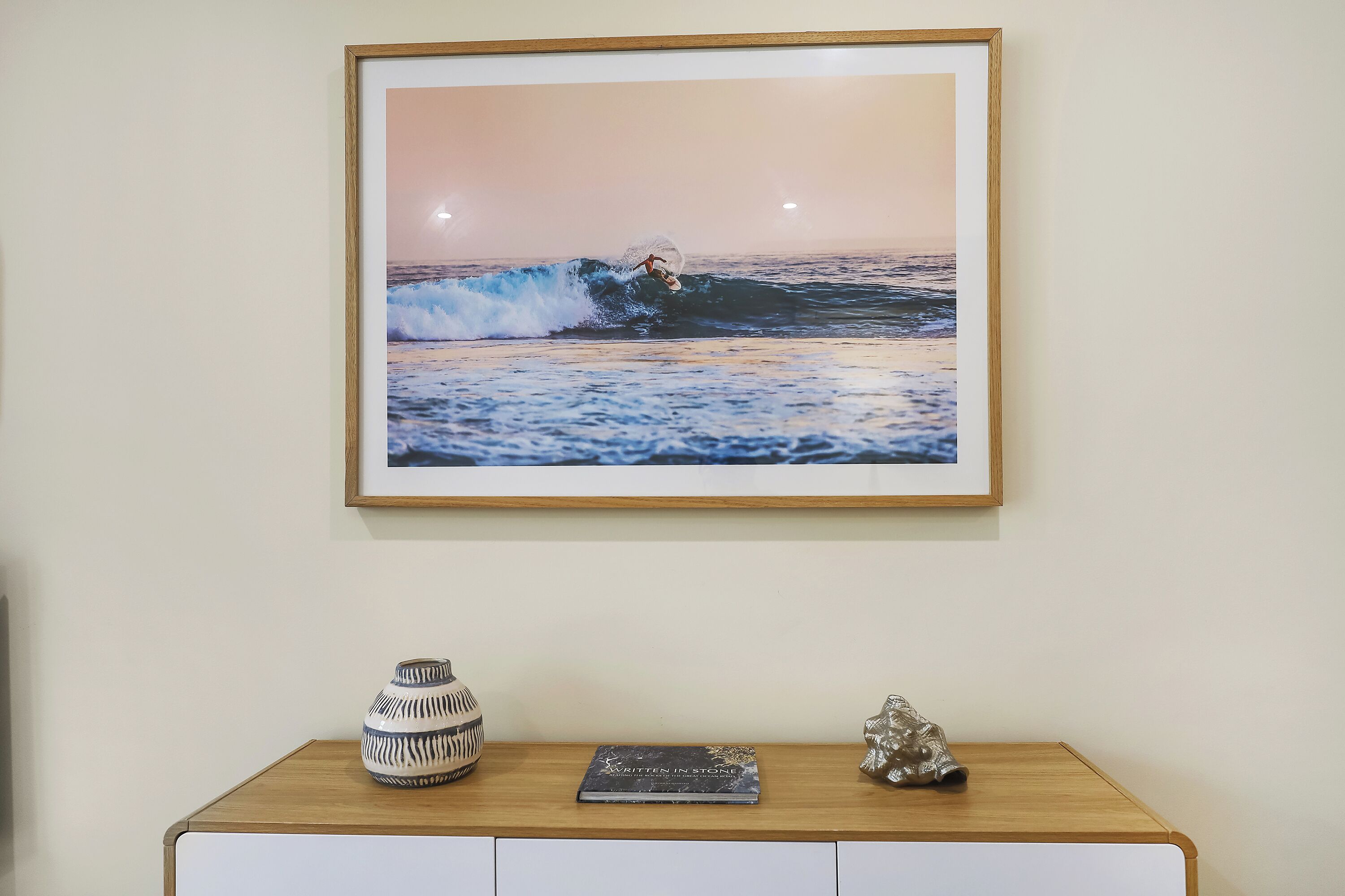 A framed photograph of a surfer on a wave, a vase, and a book on a wooden shelf.