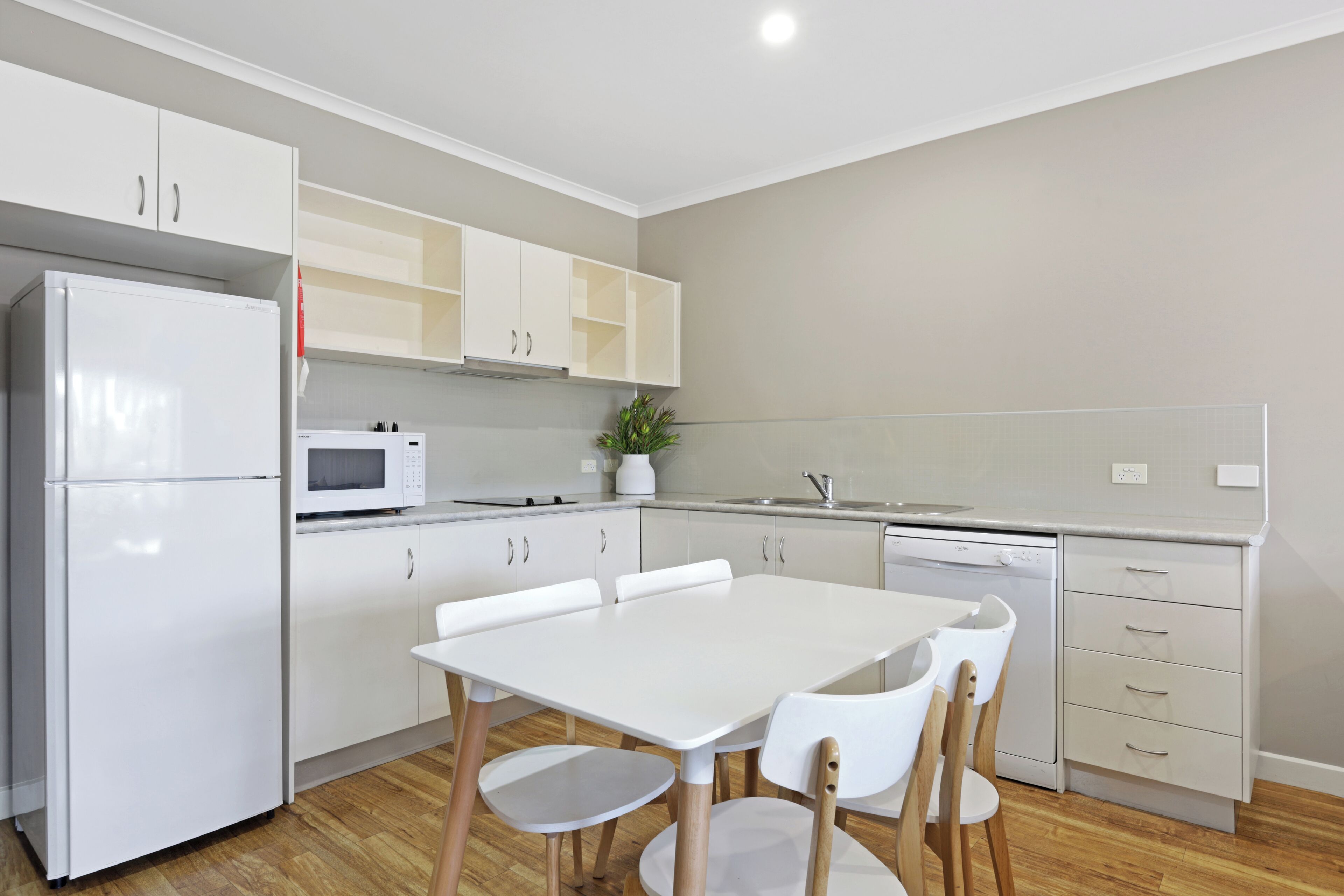 Une cuisine moderne avec des meubles blancs, un réfrigérateur blanc, un four à micro-ondes et un lave-vaisselle. On y trouve une table à manger avec quatre chaises et une plante en pot posée sur le comptoir.