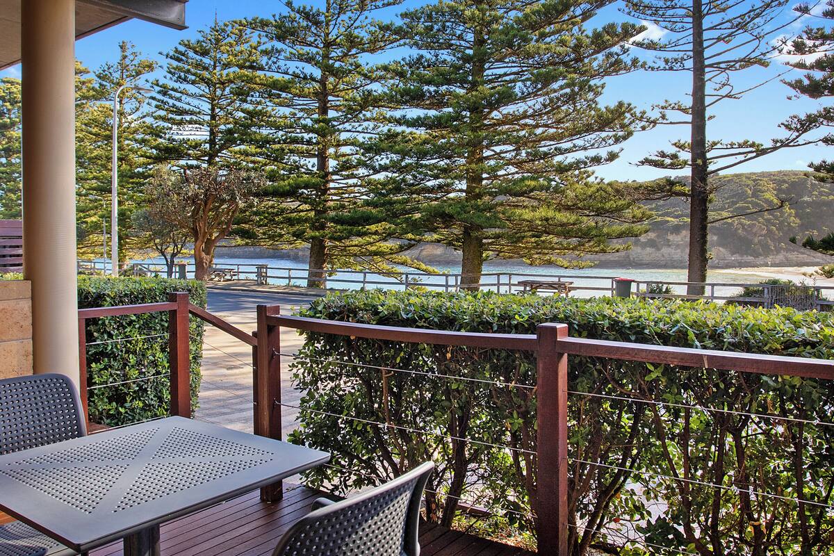 A patio with a table and chairs, overlooking a beach and trees.