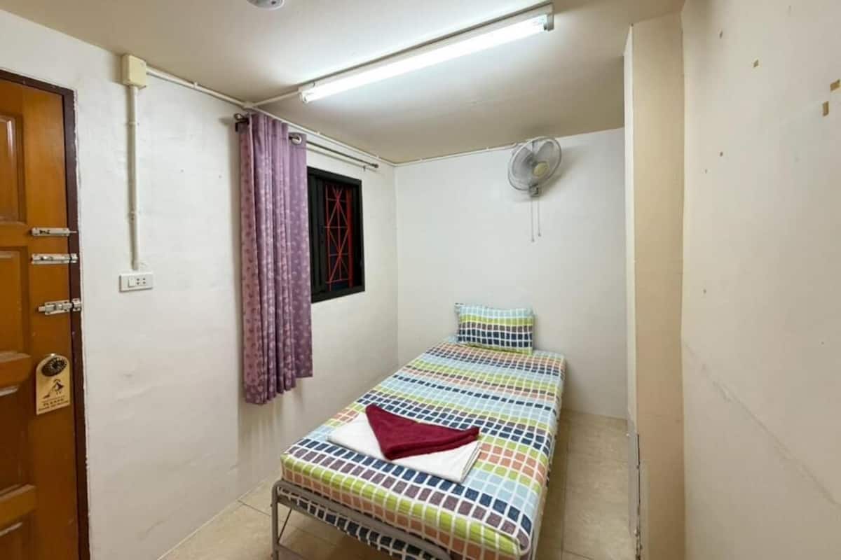 Una habitación pequeña con cama individual, ventana con cortinas, ventilador de pared y puerta con cerradura.