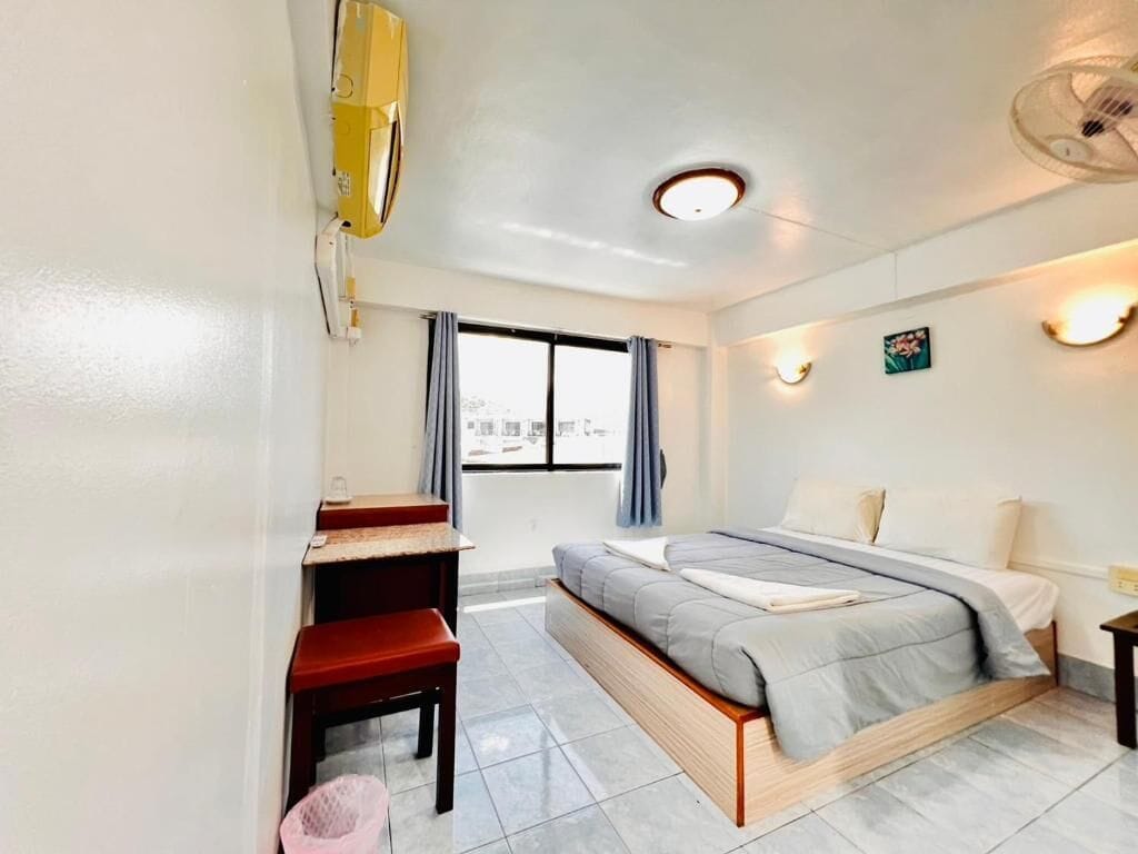 A compact hotel room with a bed, a desk, a chair, a trash bin, and a window with curtains.