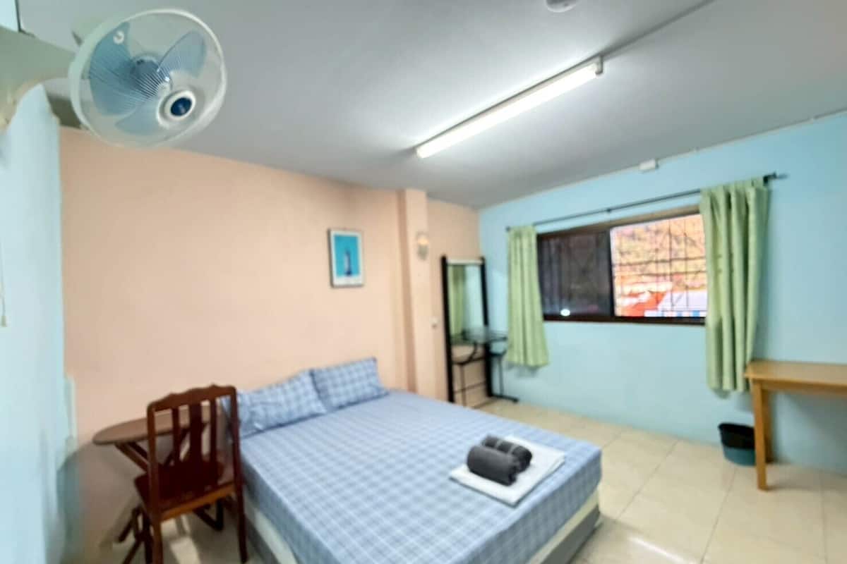 Un dormitorio con cama, una silla, una mesa, una ventana con cortinas y un ventilador de techo.