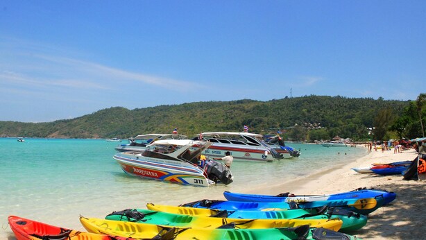 A beach with boats and kayaks, clear turquoise water, and a mountainous backdrop. - Chongkhao Resort (Ko Phi Phi)